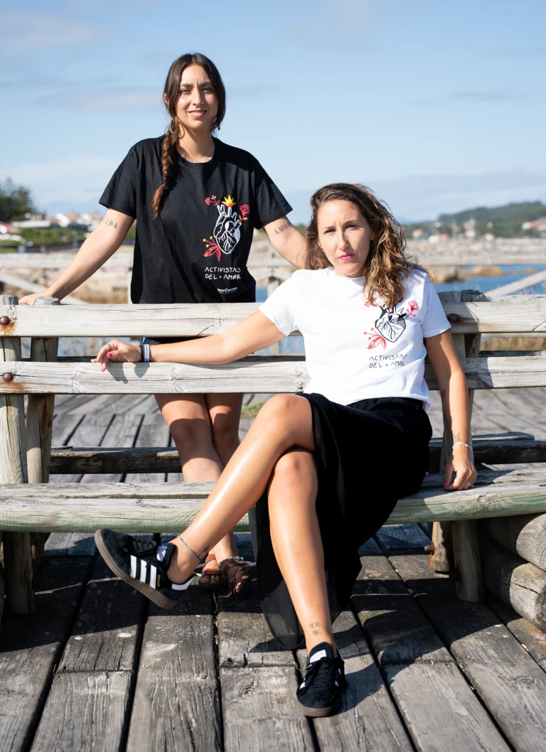 Dos activistas con camisetas de la colección Activistas del Amor, sentadas en un banco junto al mar